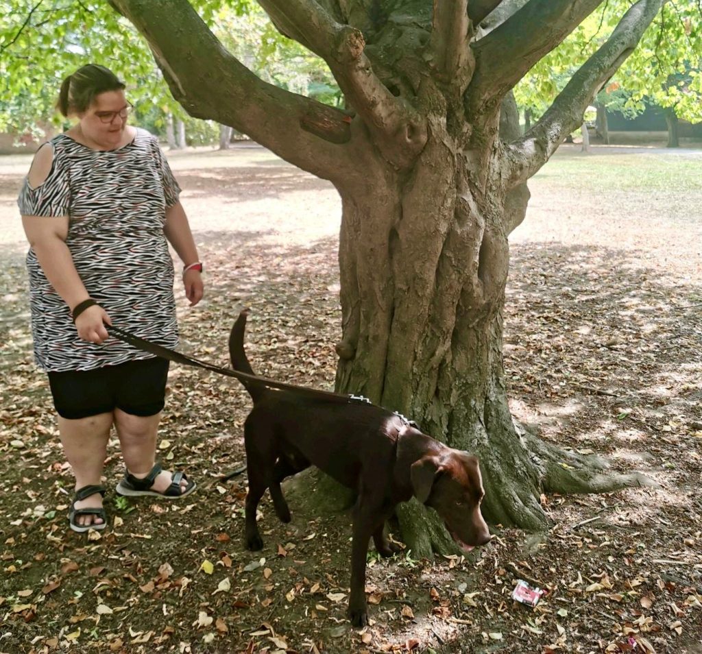 Frau steht mit Hund Baro vor einem großen Baum.