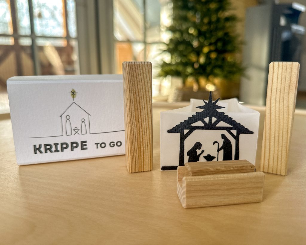 Links steht eine kleine Schachtel, darauf steht: Krippe to go. Rechts steht eine kleine Krippe aus Papier und Holz.