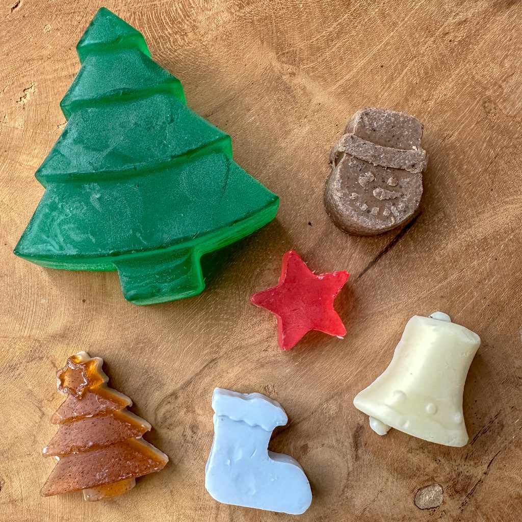 Eine grüne Tannenbaum-Seife, eine braune Schneemann-Seife, eine rote Stern-Seife, eine braune Tannenbaumseife, eine hellblaue Stiefel-Seife und eine beige Glocken-Seife.