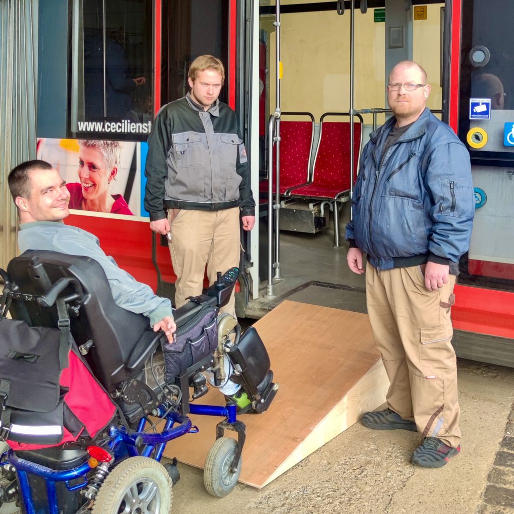 2 Mitarbeiter der Tischlergruppe und ein weiterer Mitarbeiter im Rollstuhl vor der selbstgebauten Rollstuhlrampe